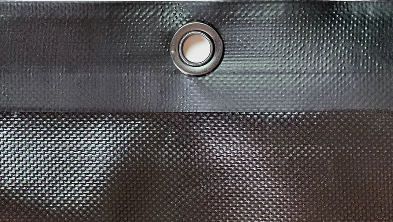 Close-up of 18 oz. vinyl PVC coated polyester dump truck tarp material showing heat-welded seam, brass grommet, and solid weave texture.